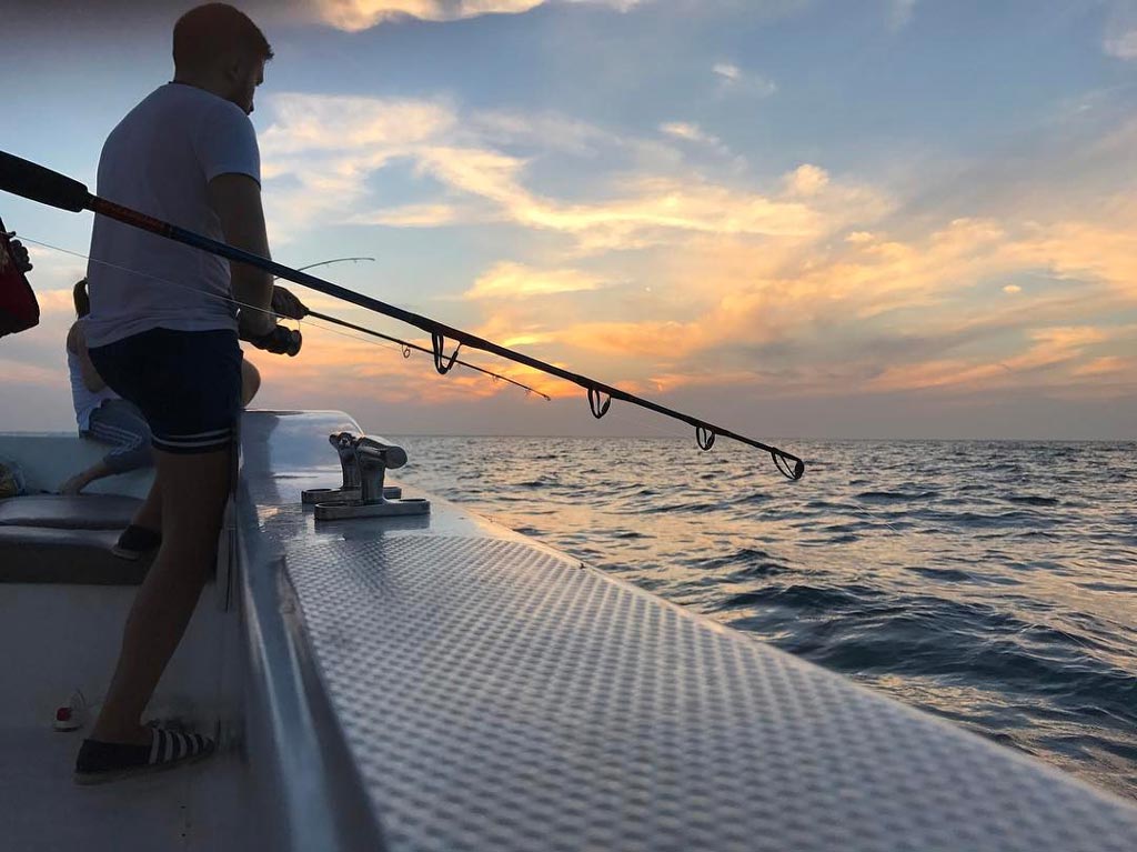 A photo taken at sunset of a fishing rod being held over the side of a charter boat, with an angler holding a second rod visible behind.