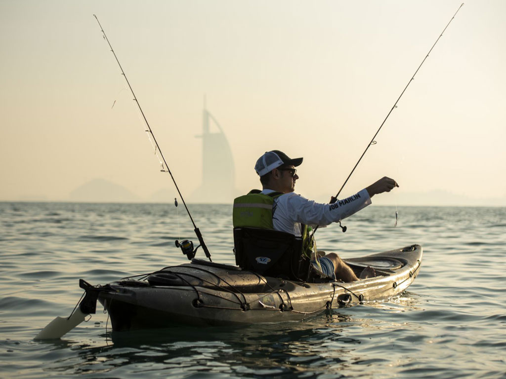 A sunset photo of an angler in a kayak, holding a lure on one of his two rods, with a silhouette of Burj Al Arab visible in the background. 