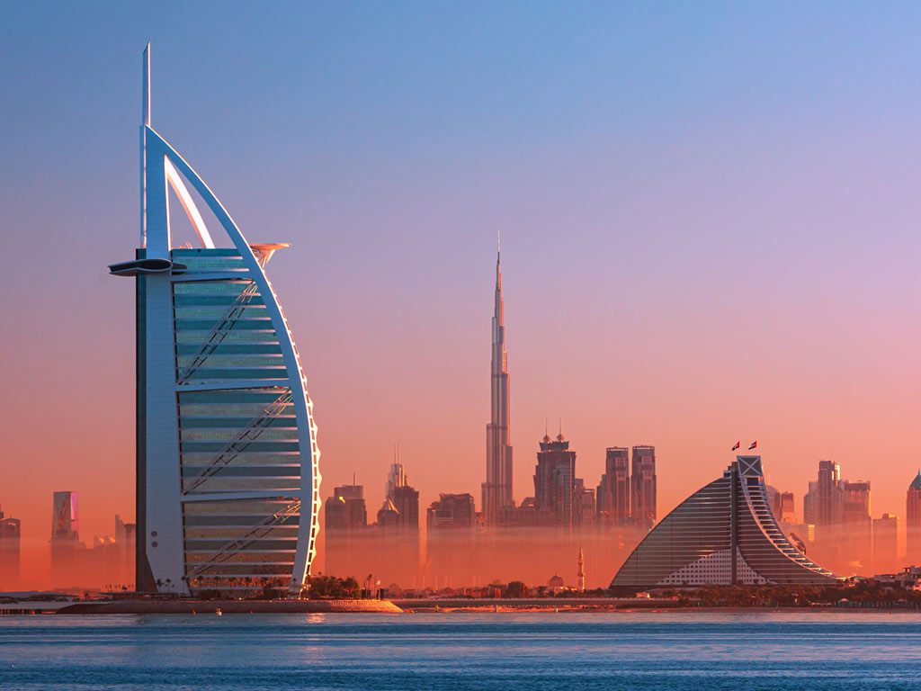 A sunset photo of Burj Al Arab and the Dubai skyline from the water, with red dust visible in the air.