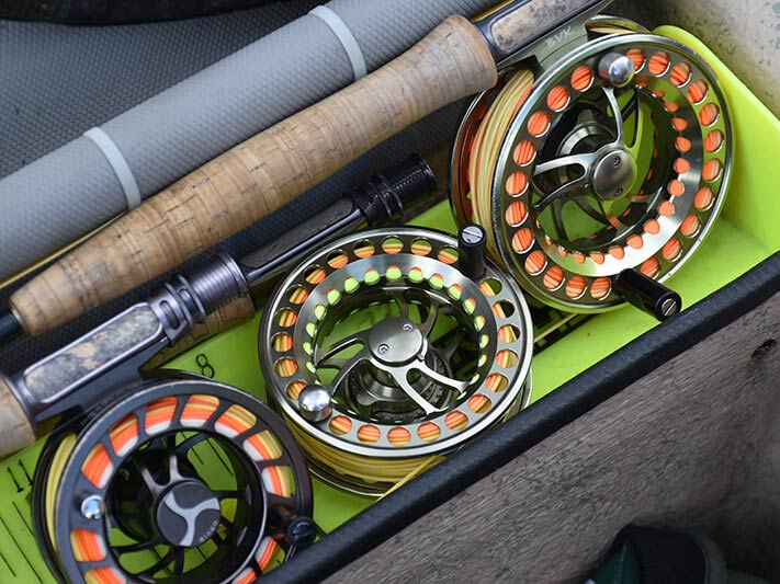 A close up of three fly fishing reels and a couple of handles of fishing rods, packed into a bag with a green bottom