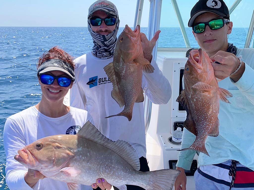 A group of smiling anglers aboard a fishing charter near Siesta Key, holding up their Grouper catches with the water behind them on a sunny day