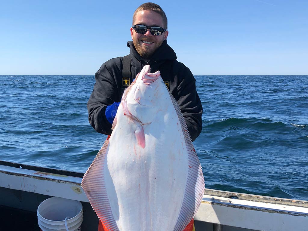 A short-haired angler wearing sunglasses, standing on a boat and holding a large Halibut with the water behind him on a clear day