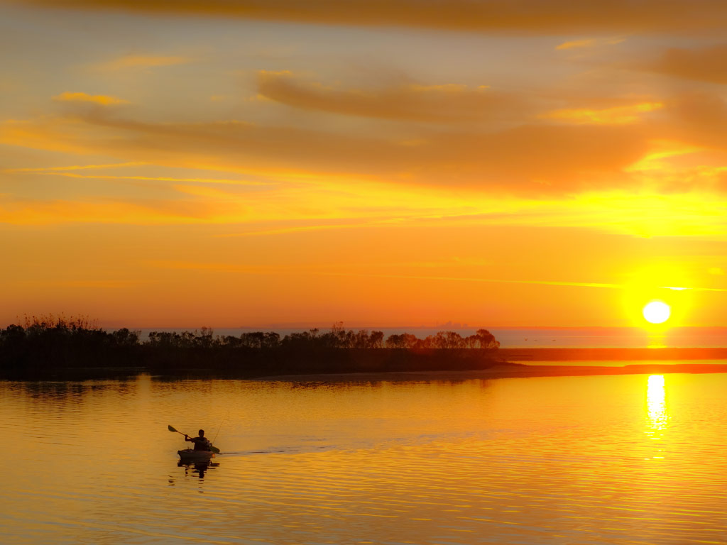 A kayaker paddles in calm waters at sunrise, with the sun casting an orange tint over the waters.