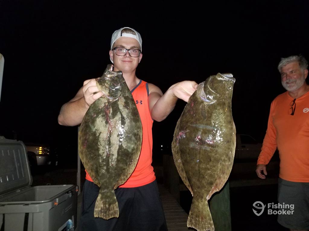 A young angler in an orange vest and a backwards baseball cap, holding up two Flounders caught on a night fishing trip near Galveston, TX, while a man in orange looks on nearby