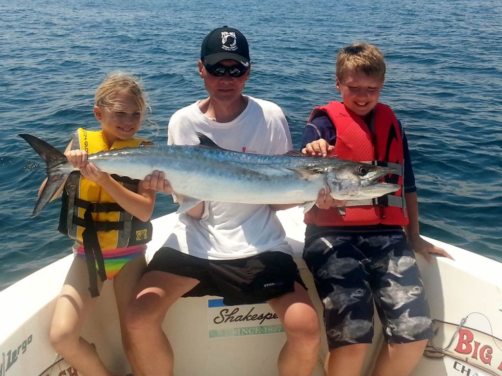 A photo of an older angler sitting on a charter boat with King Mackerel in both hands and posing with two kids in life vests standing and smiling next to the fish