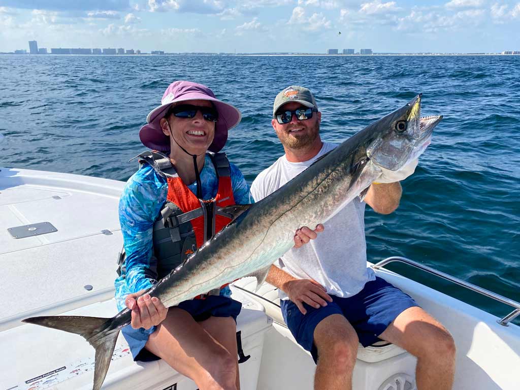 A man and a woman sitting on a charter fishing boat off the coast of Perdido Pass, both holding a big King Mackerel, with the shore visible in the distance.