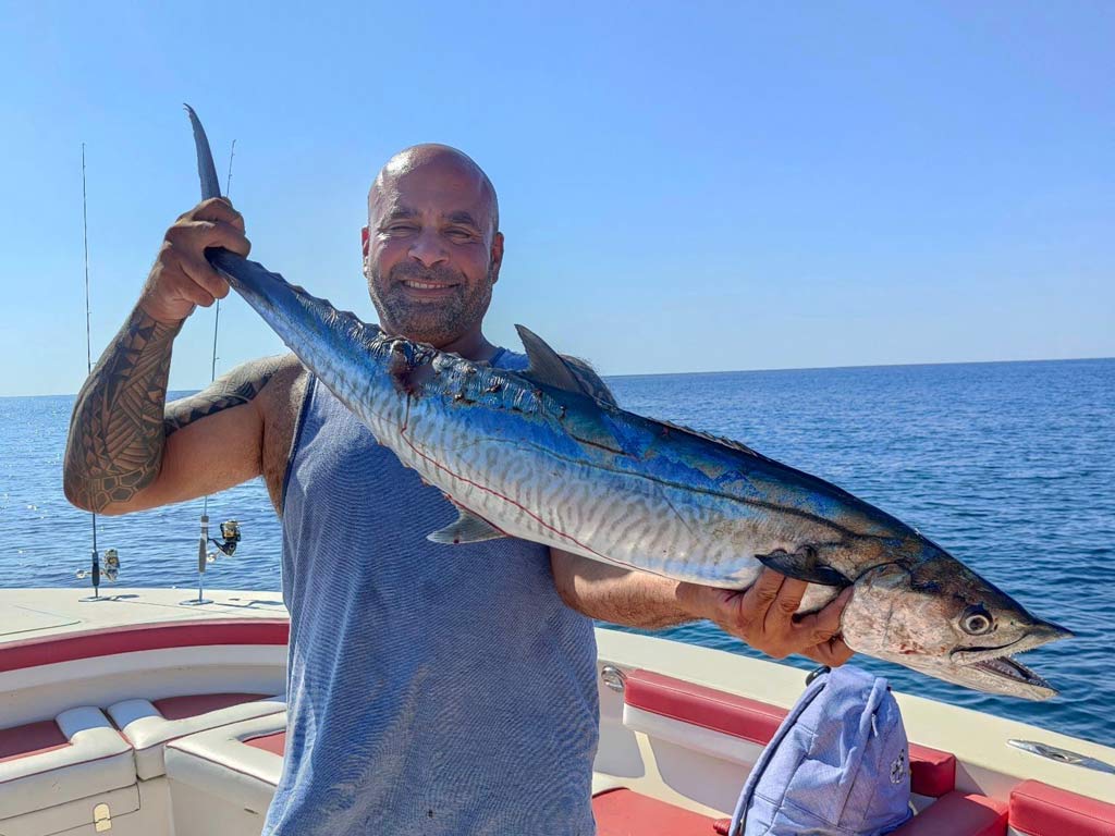 A bald man in a blue sleeveless shirt standing on a boat, holding a big King Mackerel he caught fishing in Dubai and smiling. 