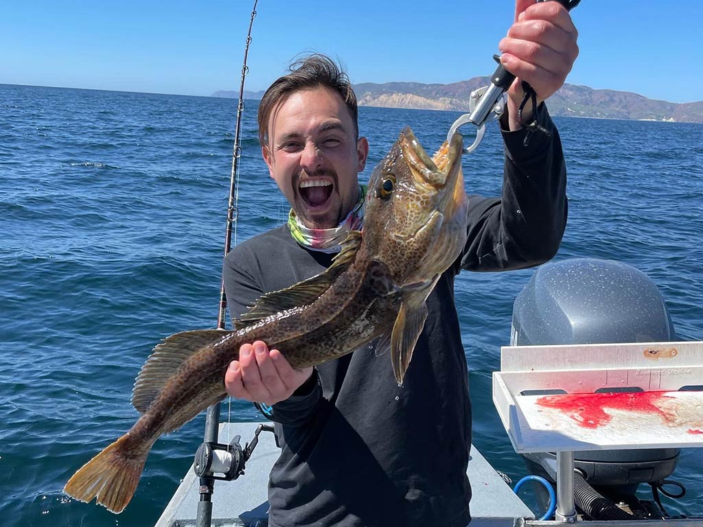 A smiling angler holding up a Lingcod he just caught to the camera, while standing on a fishing charter in California on a bright day.