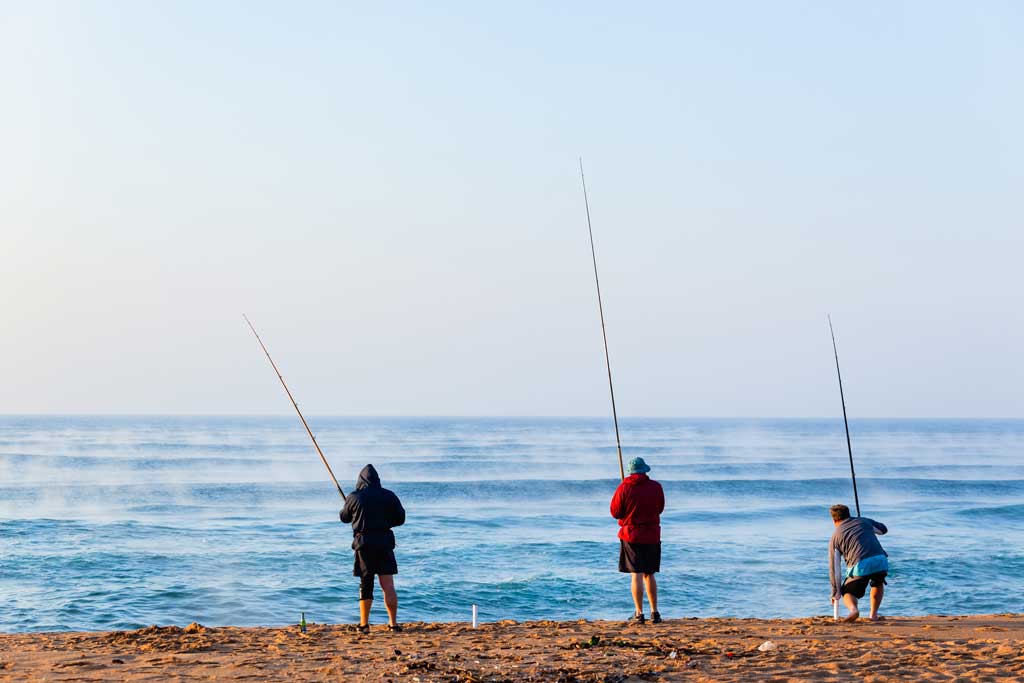 Three people surf fishing on a beach, each holding a fishing rod, with calm waters in front of them