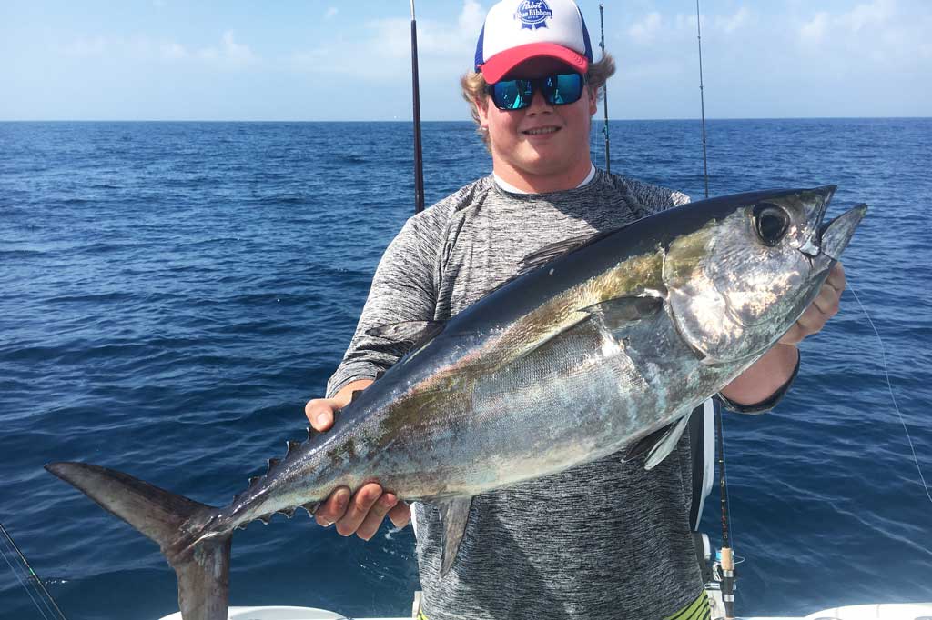 A fisherman in a cap and sunglasses, standing on a charter fishing boat, holding a Blackfin Tuna, with blue waters in the background