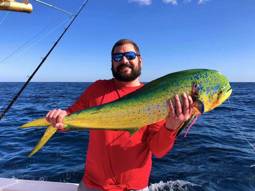 An shot of a proud angler smiling while standing on a Tybee Island charter fishing boat and holding a big Mahi Mahi caught offshore with both hands, with blue waters behind him on a clear day