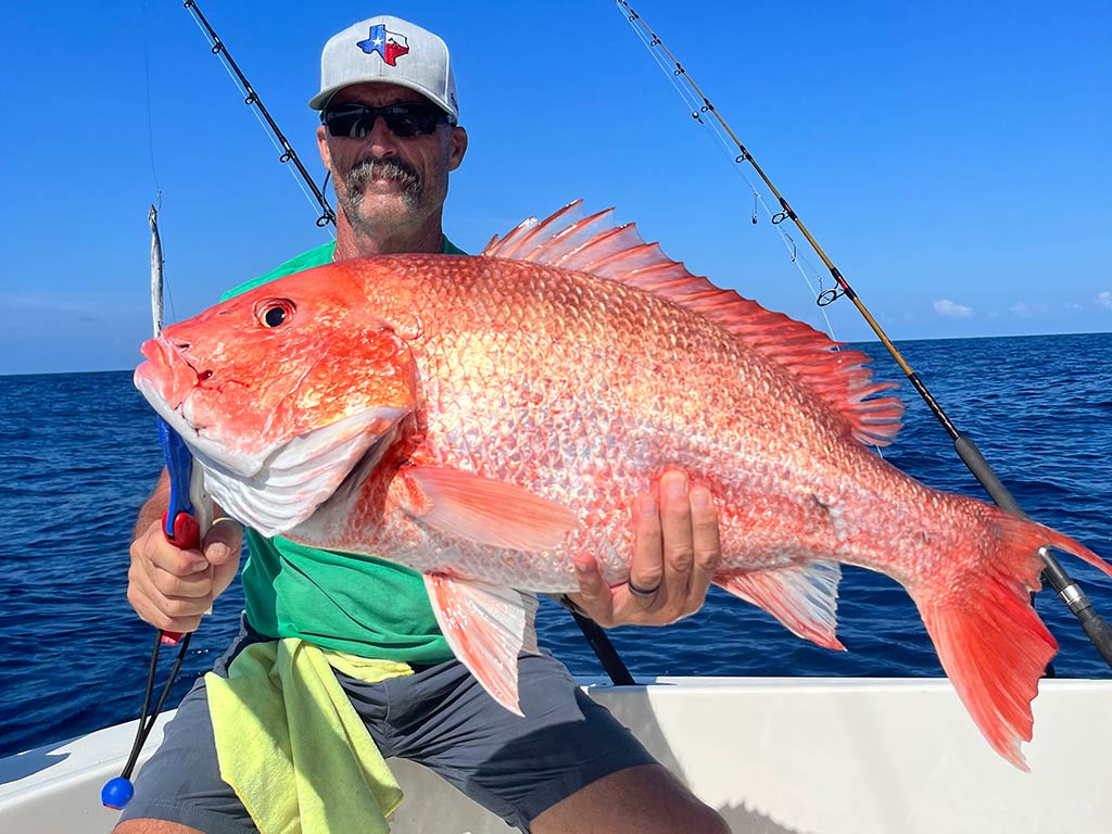 An angler with a moustache and wearing a baseball cap sits on the corner of a boat, holding a large Red Snapper on a sunny day