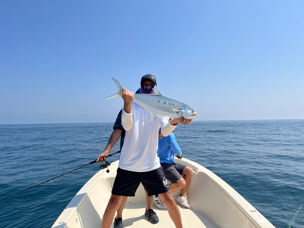 An angler on a charter fishing boat posing with a Queenfish he caught, with two other anglers partially visible behind him, as well as the ocean.