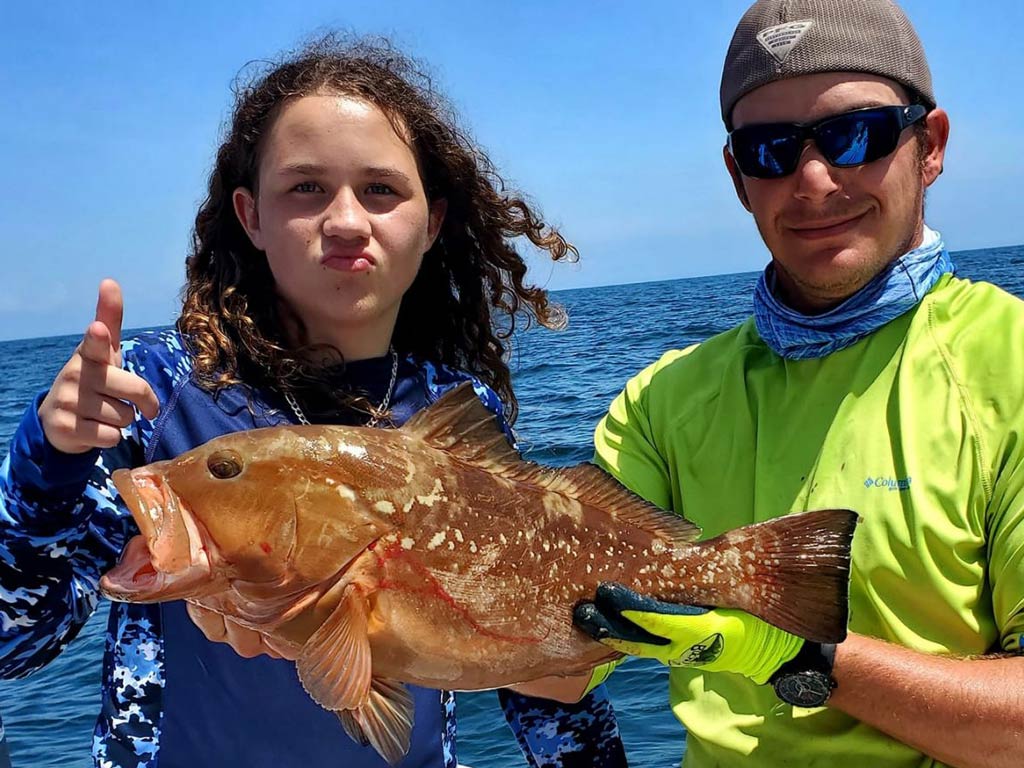 A photo of a young angler pointing to the camera next to a captain holding Grouper while posing on a charter fishing boat