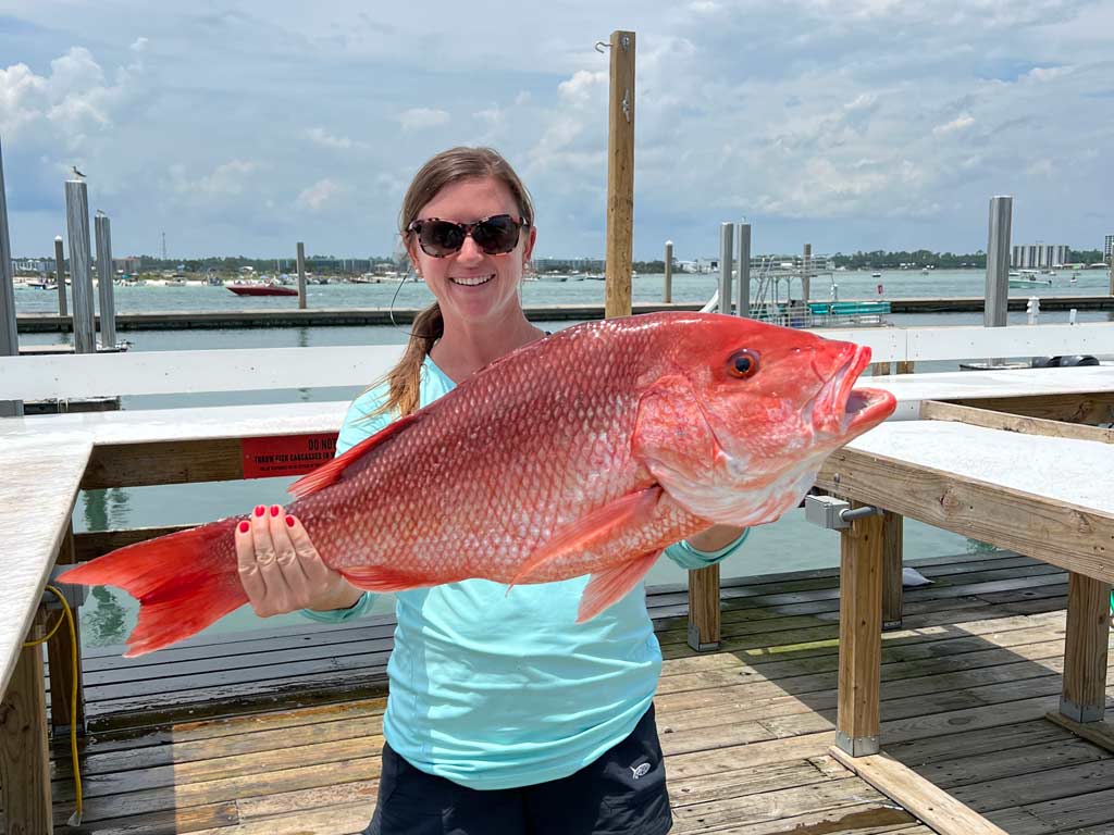 A woman in sunglasses standing on a dock in Alabama, holding a sizeable Red Snapper, with the coastal waters visible behind her.