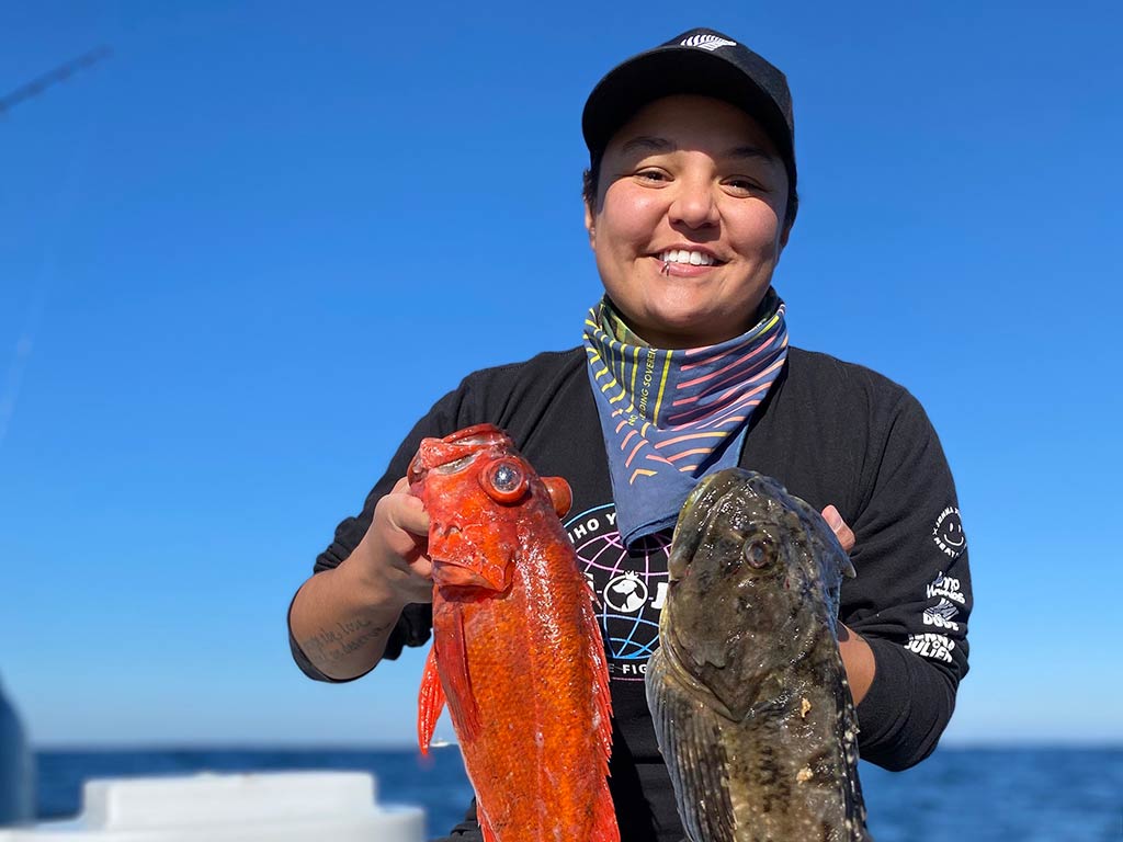 A smiling woman in a baseball cap and a neck bluff holding up two Rockfish, one orange, one brown, with clear skies behind her