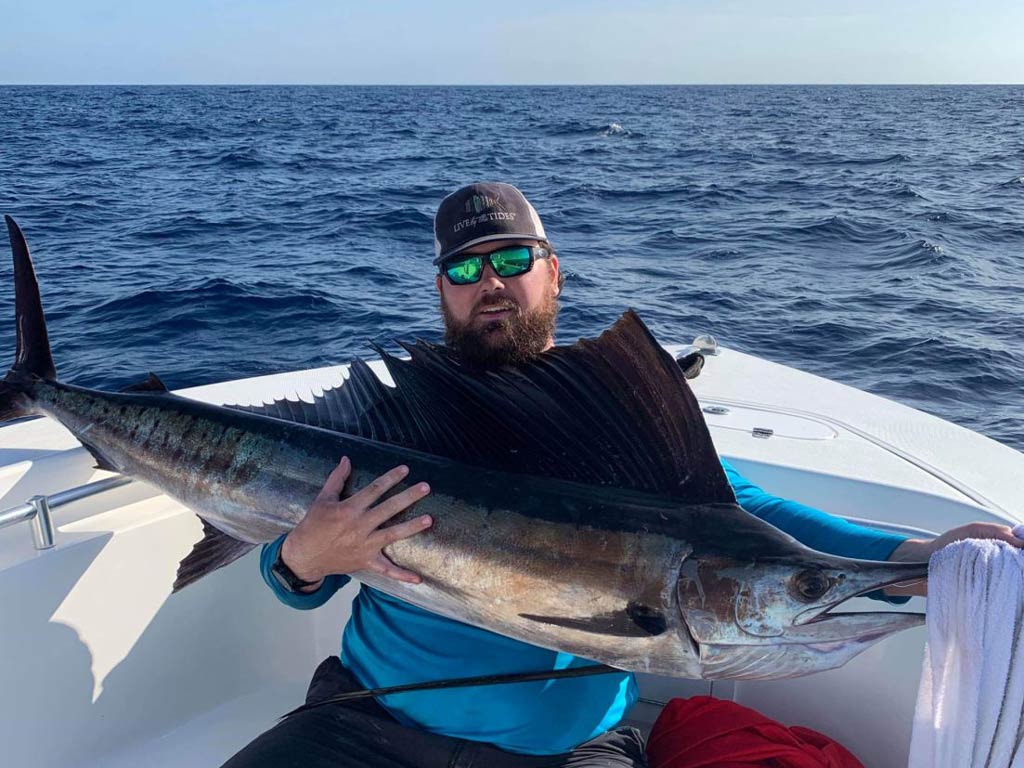 A photo of an angler wearing a cap and sunglasses while sitting on a charter fishing boat and holding a Sailfish with both hands on a bright and sunny day