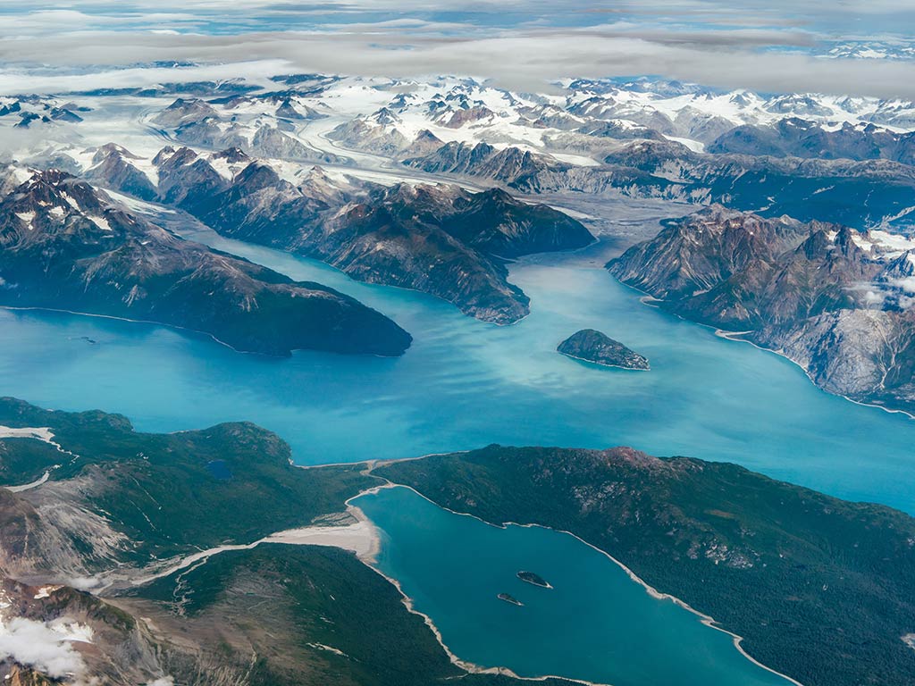 An aerial view of the fiords near Anchorage, AK, with blue eaters working their way around big mountains, some of which are snow-capped on a cloudy day