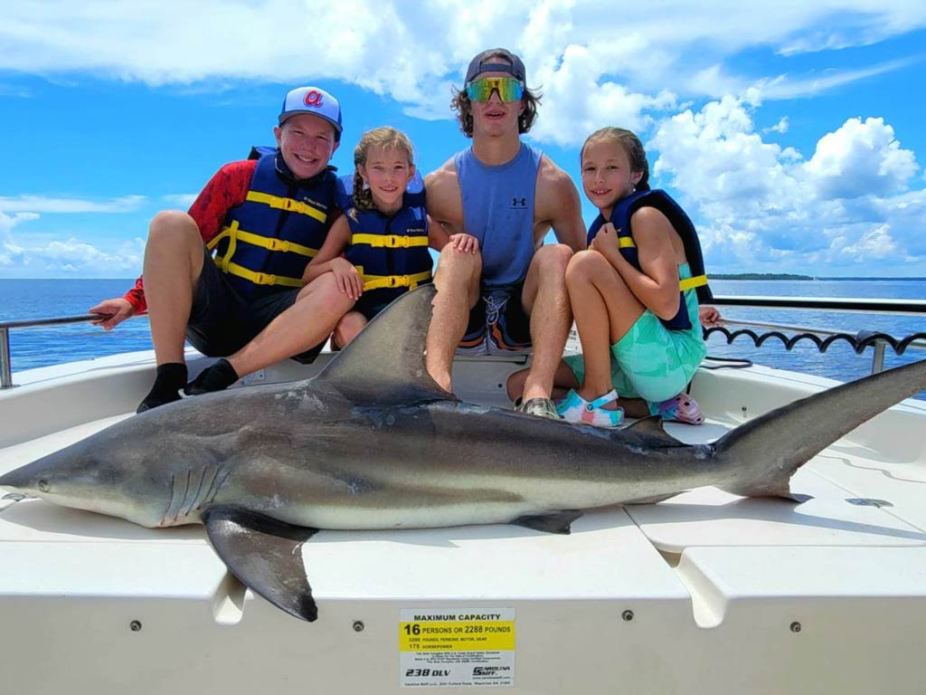 A group photo featuring three kids gathered behind a big Shark while posing for the camera on a charter boat with their captain on a bright and sunny day