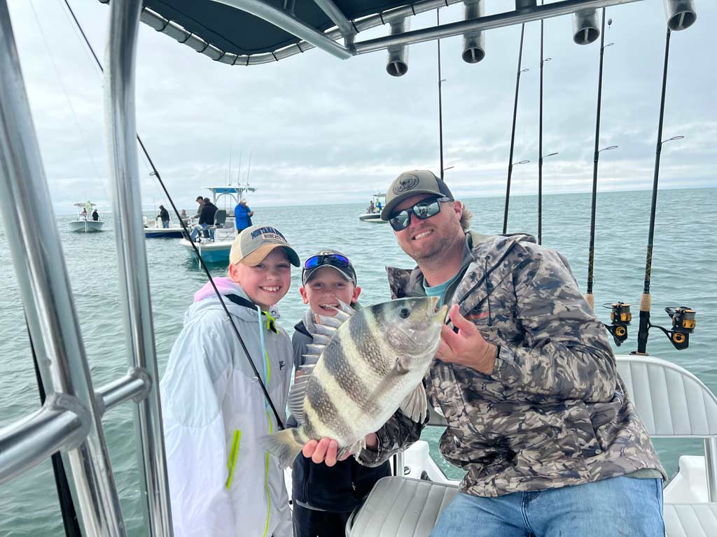 A man in sunglasses and a hat sitting in the center console seat of his boat and holding a Sheepshead he caught fishing in Perdido Pass, with two kids to the left of him posing for a photo.