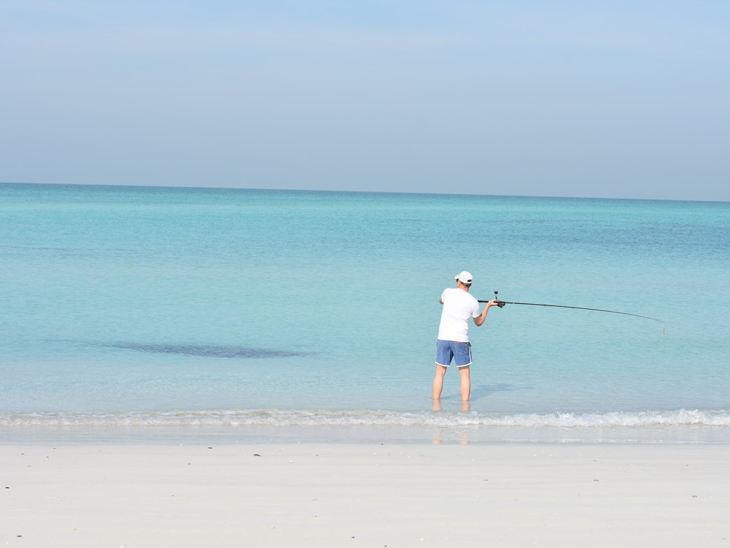 A photo of a man casting his fishing rod while ankle deep in clear, light blue waters on a white sandy beach.