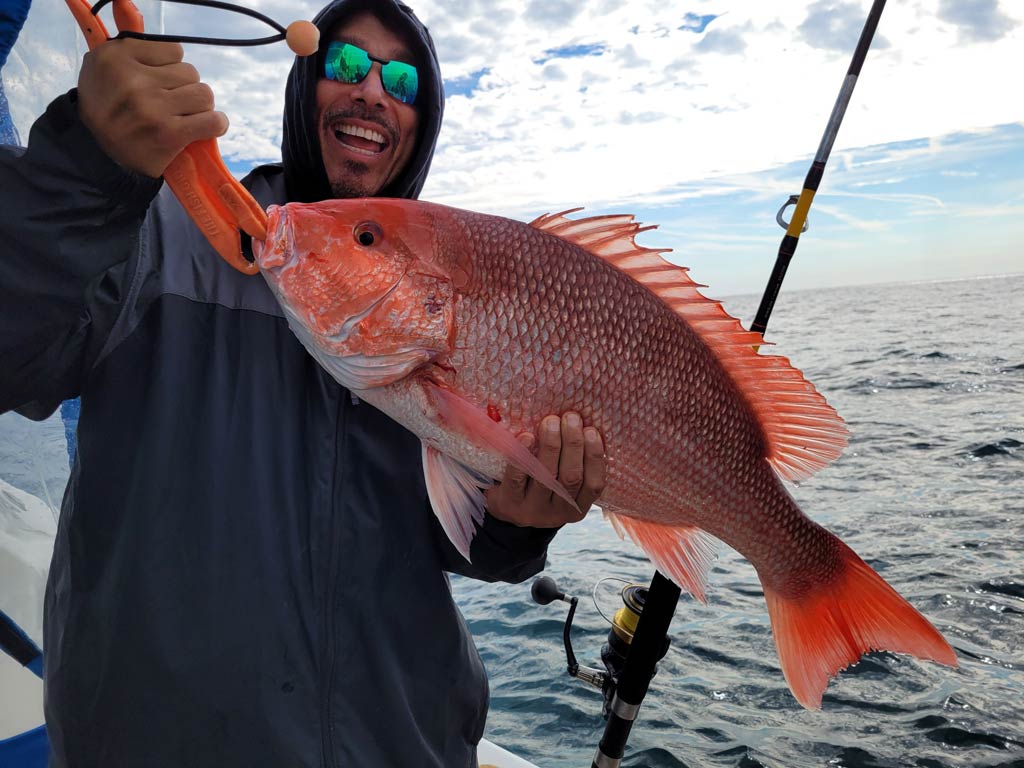A photo of a happy angler wearing a hoodie and a pair of sunglasses while standing on a charter boat and holding a big Snapper with both hands on a bottom fishing trip