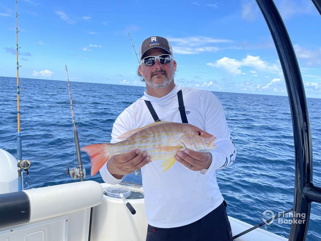 An angler in a baseball cap and sunglasses holding a small Snapper while standing on a fishing charter with the water behind him