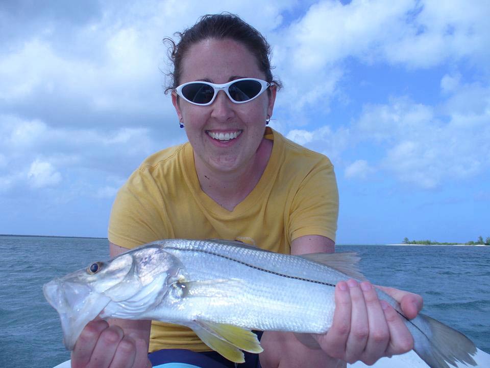 A female angler wearing sunglasses, crouching towards the camera and presenting a Snook caught while fly fishing in Cozumel on a cloudy day