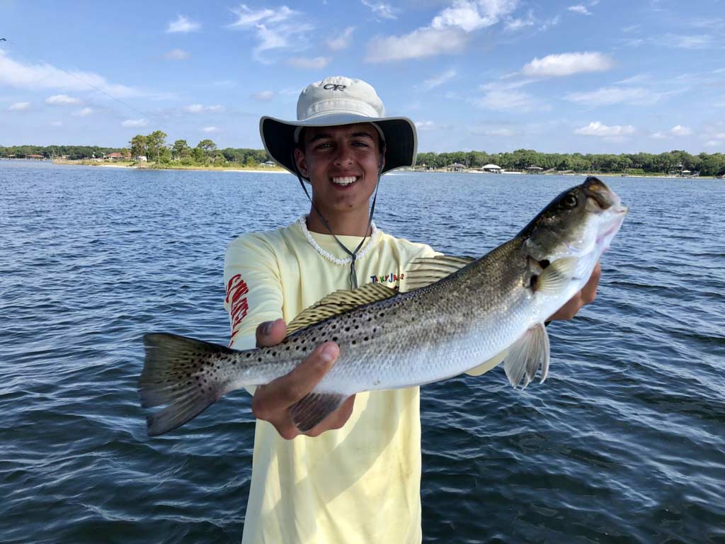 A young angler in a fishing hat holding a big Speckled Trout towards the camera with waters and the shore visible in the background.