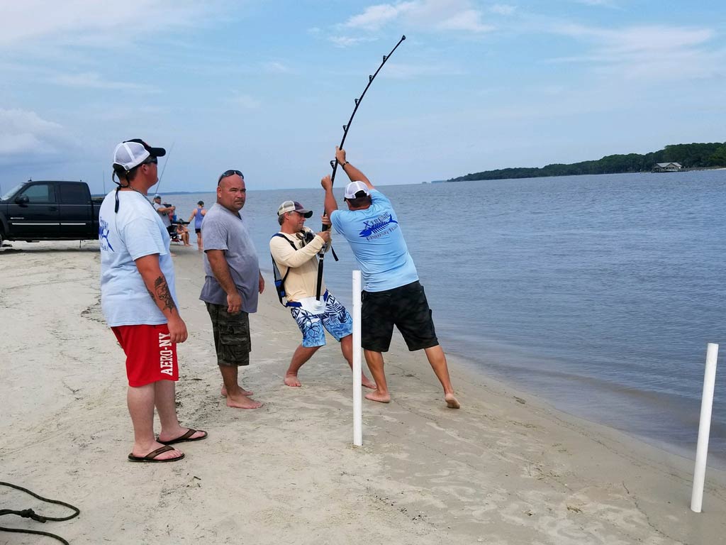 A photo featuring several surf anglers gathered around a bended fishing rod during a beach fishing trip in St. Simons Island, with two anglers struggling to reel in a fish, while the other two anglers are carefully waiting for the outcome 