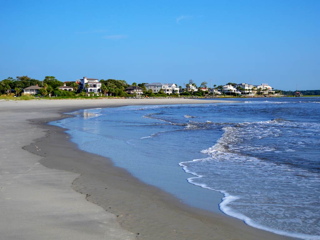 A photo featuring a sandy beach with blue waves and houses in the distance, taken on St. Simons Island on a bright and sunny day