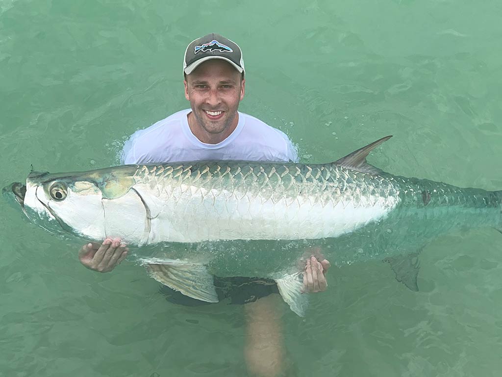 An angler up to his waist in greenish waters, holding a large Tarpon, partially submerged in the water in Siesta Key