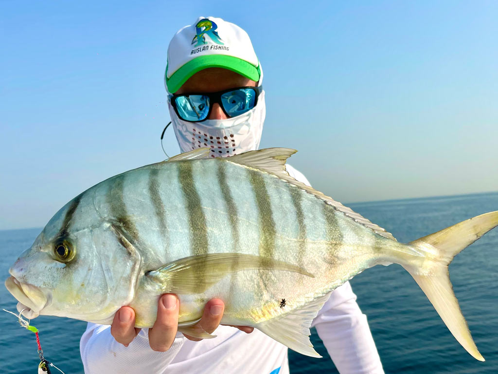 An angler in a baseball cap, sunglasses, and a neck buff holding a Golden Trevally towards the camera with one hand, with the open water behind him.