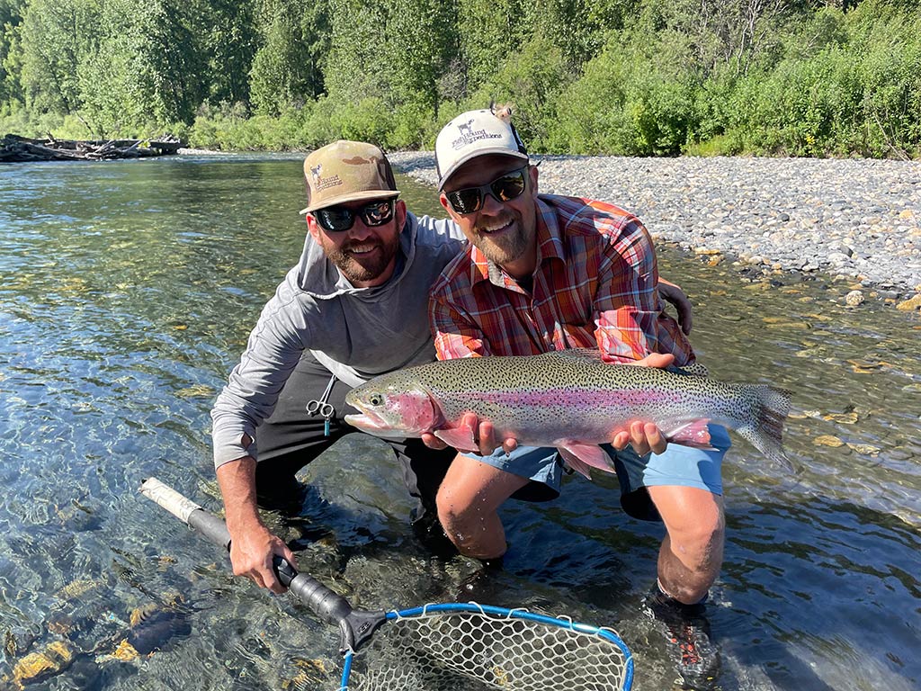 Two anglers in baseball caps crouching in shallow waters near Anchorage, AK, with one holding a large Rainbow Trout, while the other holds a fishing net on a sunny day