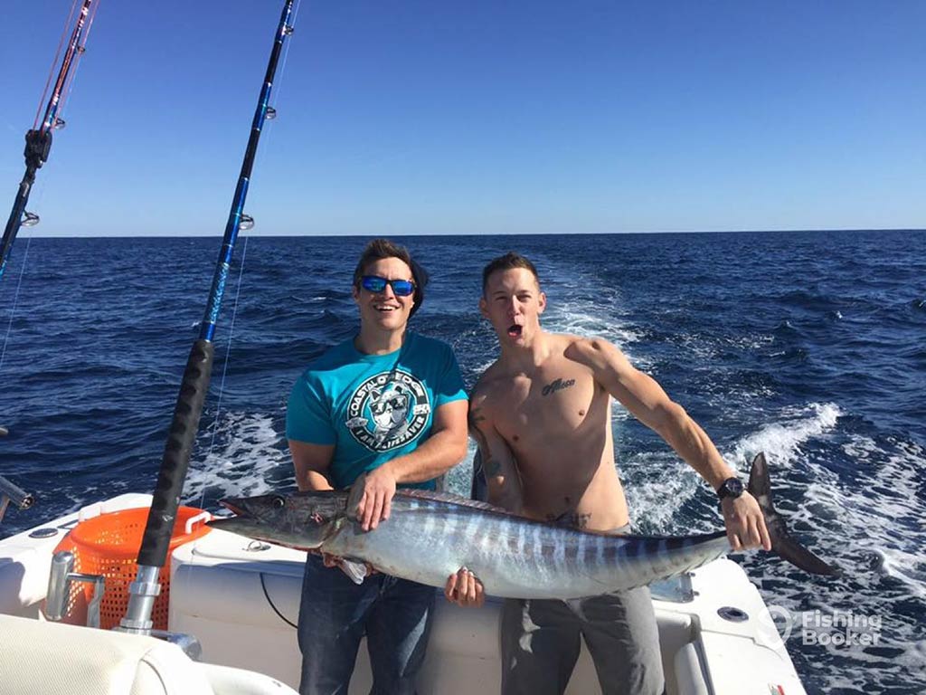 Two anglers, one of them shirtless, sitting on the deck of a boat and holding a large Wahoo offshore from Georgetown, SC, with the wake of the boat behind them on a sunny day