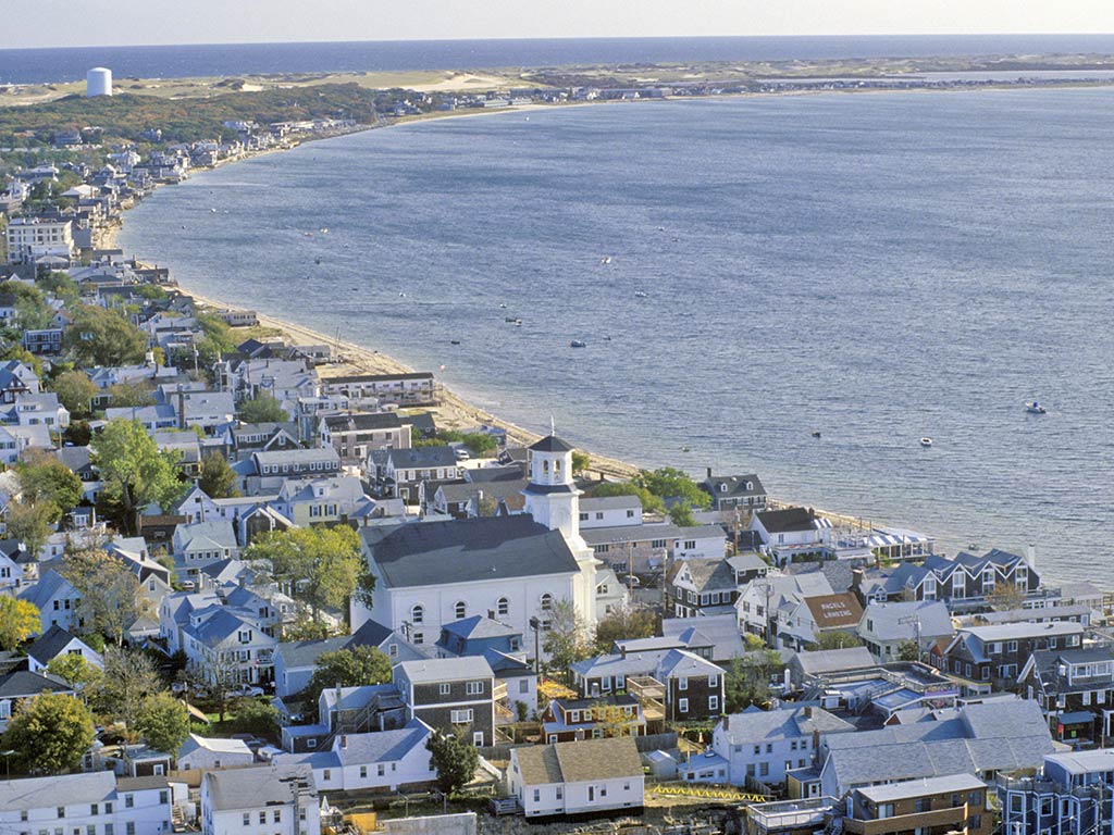 An aerial view of Provincetown, MA, with the Atlantic Ocean on the right-hand side of the image and white buildings including a large church in the foreground