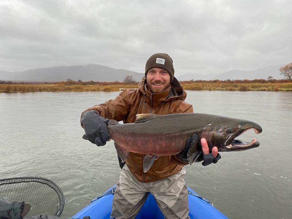 An angler wearing a hat and a winter jacket holding a large Arctic Char while stood on a fishing boat near Anchorage on a cloudy day with the water behind him