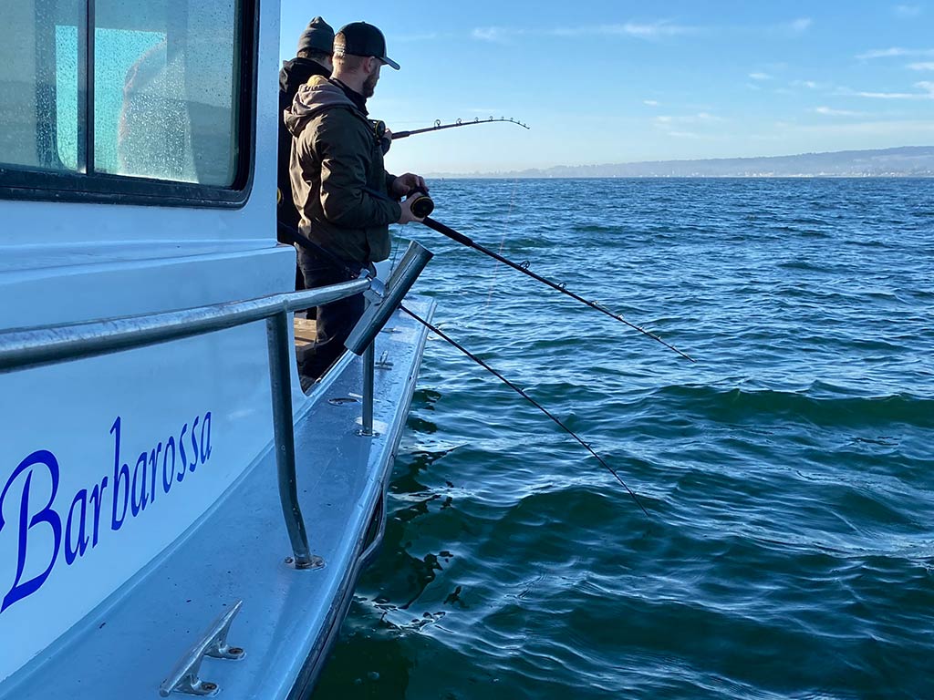 A view along the side of a boat of two anglers fishing out of Santa Cruz, CA on a clear day, with their lines in the water