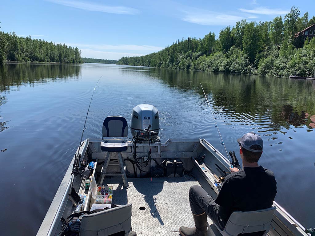 A view towards the back of a fishing boat of a river in Alaska on a clear day, with a man sitting and holding a rod while fishing, and another rod placed on the other side of the boat with a line in the water