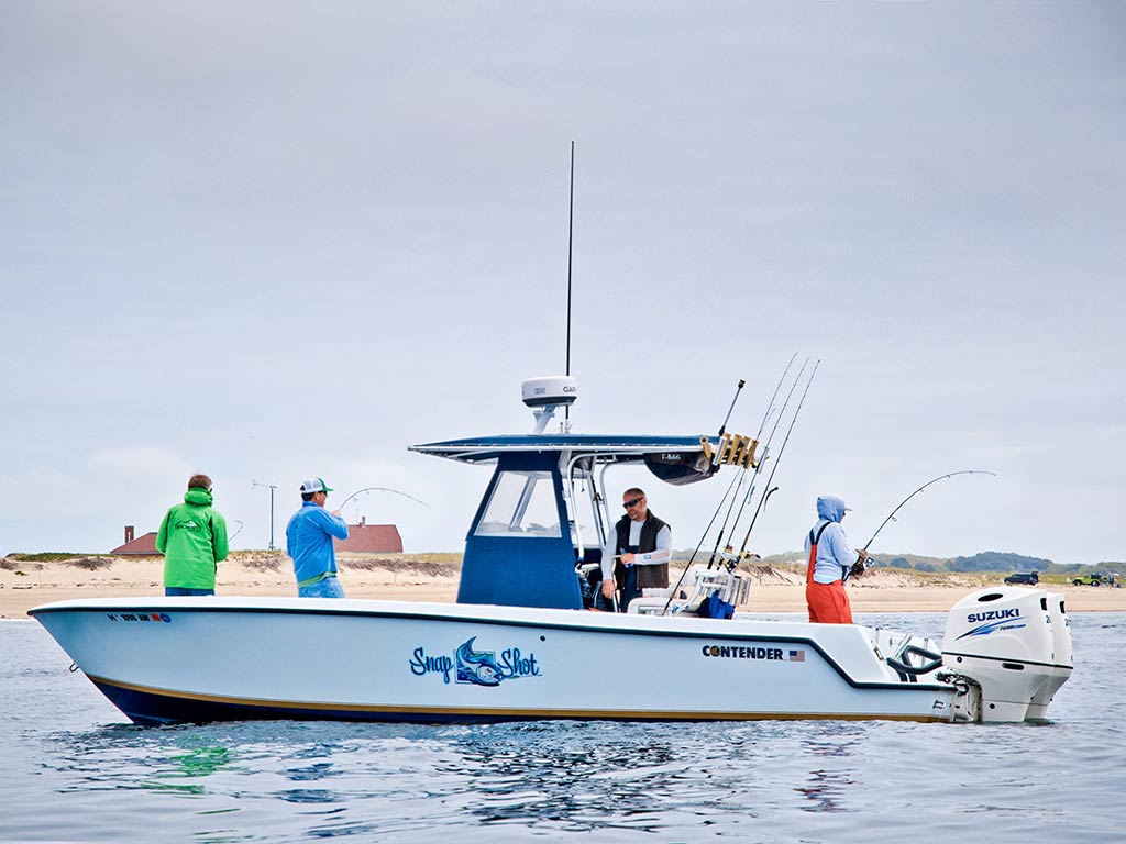 A view across the water towards a center console fishing charter with three anglers casting a line into the water from the boat, near the shore in Gloucester, MA