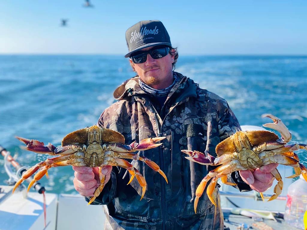 An angler in a baseball cap, stood on a fishing charter and holding two Dungeness Crabs, with the water behind him on a clear day