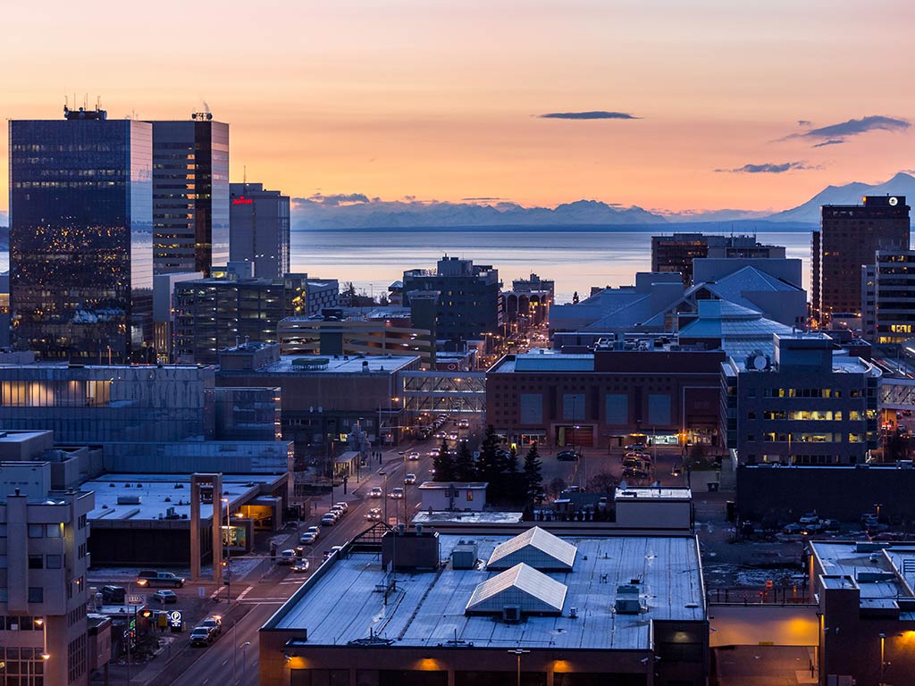 A view from high up in anchorage across the cityscape towards the water at sunset, with orange-pink skies in the distance