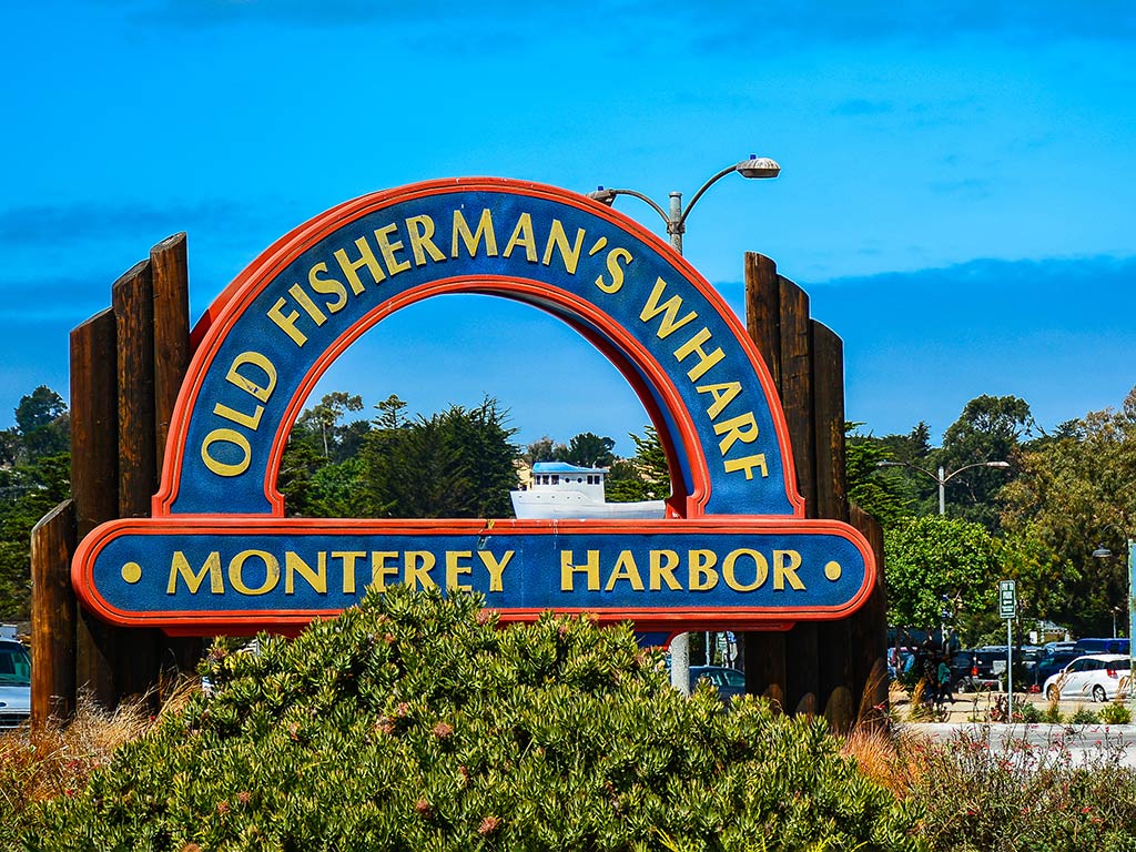 A round blue and red sign with yellow lettering saying "Old Fisherman's Wharf: Monterrey Harbor" behind some bushes on a clear day