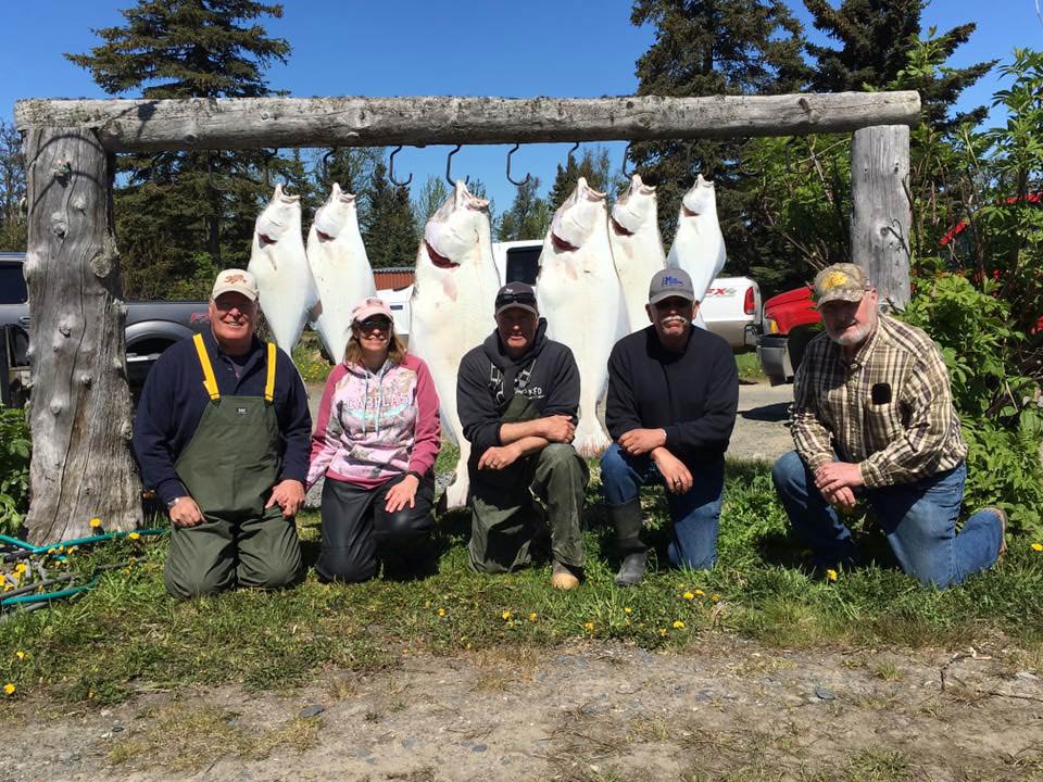 A group of anglers crouch down on the crass in front of a wooden square featuring hooks from which are hanging a number of large Halibut, caught near Anchorage