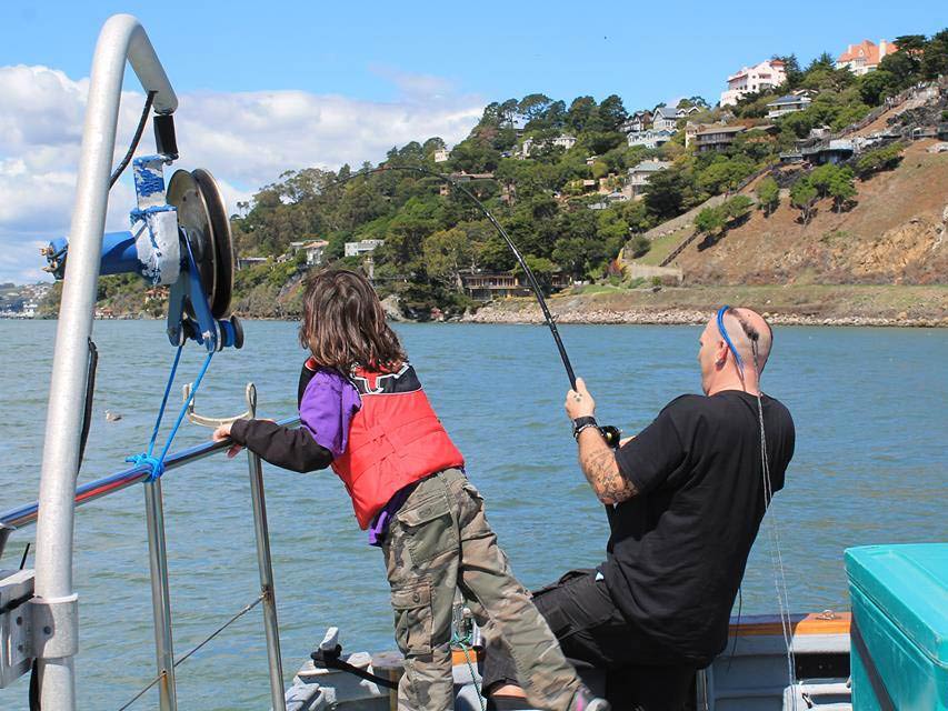 A view from behind of a balding angler sitting on the side of a boat, battling it out with a fish on the end of his line, with a child to his left looking at the water on a sunny day in California