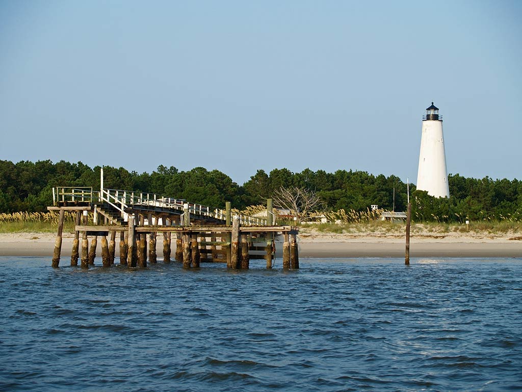 A view of a small fishing pier in Georgetown, SC, as seen from the water, with a lighthouse behind it and to its right on a calm, clear day