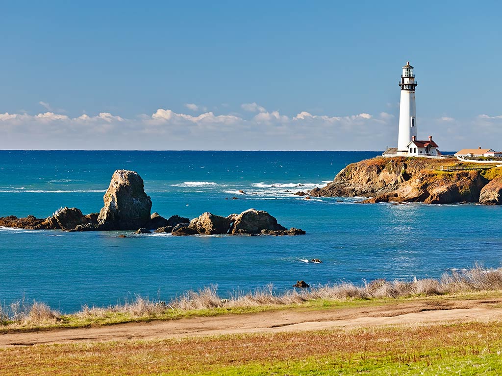 A view across a grassy beach towards some rocks in the Pacific Ocean and a lighthouse in the distance in Monterey, CA