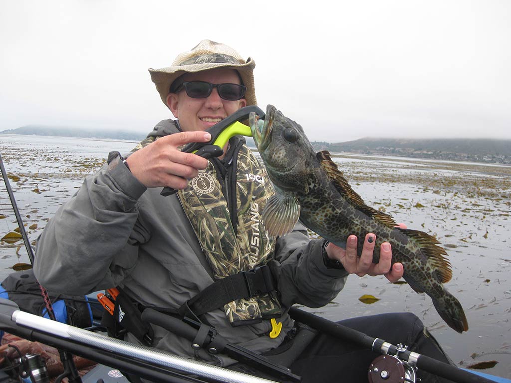 An angler in a kayak, holding up a green Lingcod to the camera, with murky waters behind him on a cloudy day