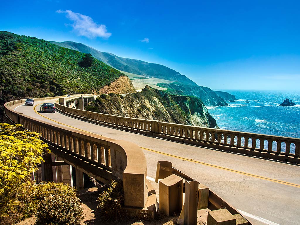 A view across a road bridge in Monterey, CA, with the ocean on the right-hand side of the image and mountains and cliffs on the left and in the distance on a clear day