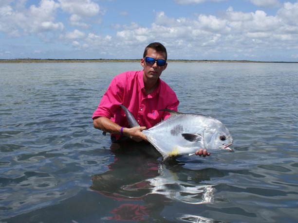 An angler in a red-pink shirt, wading up to his waist and holding a large Permit, caught fly fishing in Cozumel